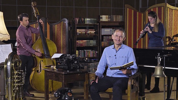 Ein Mann sitzt mit einem Buch im Sessel, im Hintergrund ein Mann mit Kontrabass und eine Frau mit Oboe.