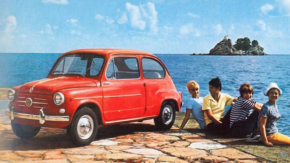 Roter Kleinstwagen am Meer vor einer kleinen Insel und vier Leute sitzen daneben mit den Beinen im Wasser.