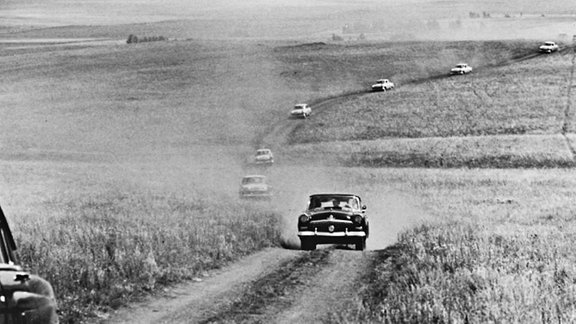 Autokolonne fährt auf gewundener Straße durch eine weite Feldlandschaft