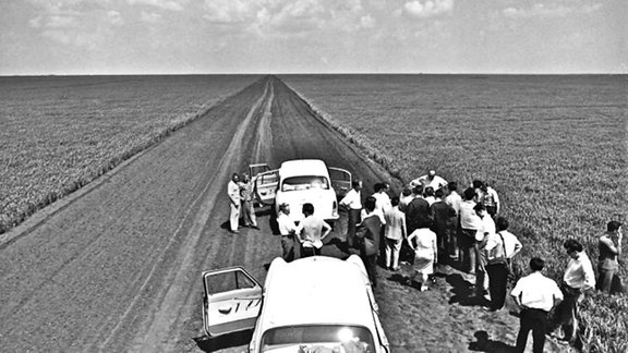 schnurgerade Straße in einem schier endlosen Feld. Zwei weiße Autos am unteren Bildrand, Frauen und Männer in Kostümen und Anzügen bestaunen die Weite der Landschaft