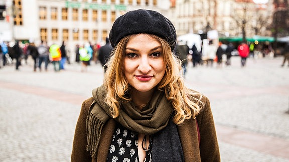 Eine junge Frau mit schulterlangem Haar und Baskenmütze auf einem Marktplatz.