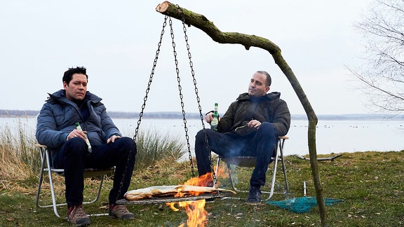 Zwei Angler sitzen an einem See in Campingstühlen am Feuer