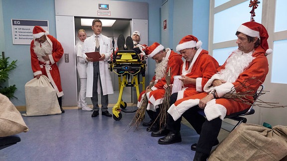 Dr. Rolf Kaminski (Udo Schenk, mi.) steht in der Notaufnahme. Im Raum sitzen vier verkleidete Weihnachtsmänner, die sich die Bäuche halten. 