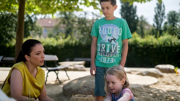 Arzu Ritter (Arzu Bazman, li.) ist mit ihren beiden Kindern Oskar (Leonard Scholz, oben,) und Pauline (Lara Schierack, unten) auf dem Spielplatz. Arzu ist mit ihren Kindern Oskar und Pauline auf dem Spielplatz. Oskar steht hinter seiner Schwester, die mehrere Sandkuchen gebacken hat.