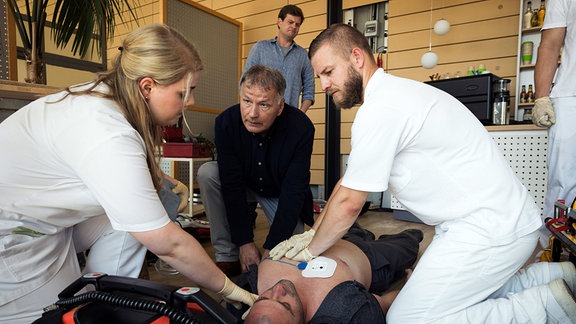 Mehrere Menschen, darunter Dr. Heilmann, knien neben dem auf dem Rücken liegenden Elektriker, der verletzt am Boden liegt.