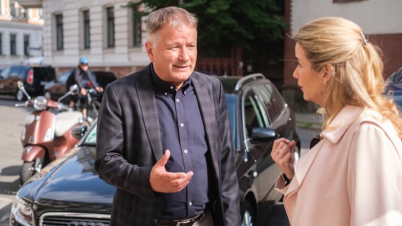 Roland und Sarah stehen vor einem schwarzen Auto. Sarah ist sichtlich überrascht, denn Roland will sie abholen.