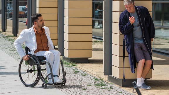 Ilay fährt in seinem Rollstuhl auf einen Mann zu, der an einer Mauer lehnt. Der Mann trägt einen dunklen Bademantel.