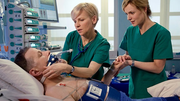 Dr. Kathrin Globisch (Andrea Kathrin Loewig, li.) und Dr. Lea Peters (Anja Nejarri, re.) leiten die Aufwachphase ein. Nun wird sich zeigen, ob Dr. Roland Heilmanns (Thomas Rühmann, liegend) Hirn Schaden genommen hat, nachdem sein Kreislauf 45 Minuten stillstand