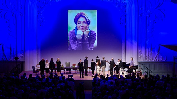 Ein Bild von Hendrikje Fitz wird eingeblendet, davor das Ensemble von In aller Freundschaft auf der Bühne