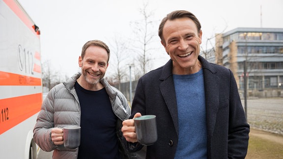 Julian Weigend und Bernhard Bettermann stehen mit Kaffetassen vor der Sachsenklinik