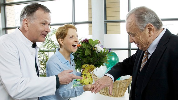 Pia Heilmann (Hendrikje Fitz, mi.) ist die neue Pächterin der Physiotherapie der Sachsenklinik. Zur Neueröffnung ist auch Professor Simoni (Dieter Bellmann, re.) unter den Gästen. Er will seinen Besuch nutzen und sich mal wieder gründlich durchchecken lassen. Doch vorher werden Neuigkeiten ausgetauscht: Pia und Roland Heilmann (Thomas Rühmann, li.) freuen sich sehr, als der Professor seinen Ehering präsentiert.