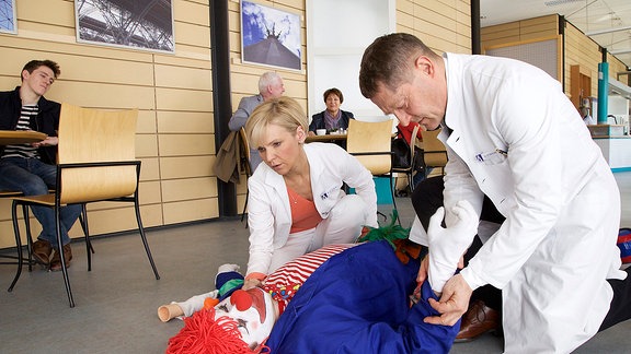 Klinikclown Abaro (Michael Schönemann, liegend) ist vom Abariskonzern geschickt worden, um unter den Patienten und Mitarbeitern für gute Laune zu sorgen. Doch gleich bei seinem ersten Auftritt in der Cafeteria bricht er vor den Augen von Dr. Kathrin Globisch (Andrea Kathrin Loewig, li.) und Dr. Kaminski (Udo Schenk, re.) zusammen.