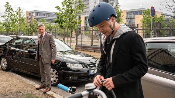 Kris steht an seinem Fahrrad, den Blick gesenkt. Hinter ihm steht Dr. Kaminski in einem schwarzen Mantel und schaut ihn böse an.