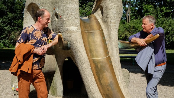 Zwei Männer stehen sich auf einem Spielplatz an einer Rutsche mit dem Aussehen eines Elefanten gegenüber und sprechen miteinander.