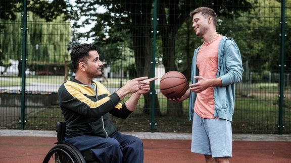 Dr. Demir und Pfleger Kris sitzen und stehen auf dem Basketballplatz und grinsen einander an.