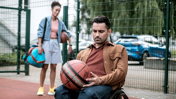 Dr. Demir sitzt in seinem Rollstuhl und hält einen Basketball in Händen. Hinter ihm steht seine Patientin.