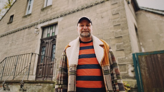 Ein bärtiger Mann mit gestreiftem Pulli, Fleecejacke und Basecap steht vor einem alten Landhaus.