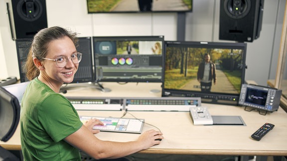 Eine junge Frau steht an einem Stehpult, auf dem Bildschirme, Mischpult und eine Tastatur stehen.