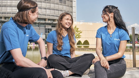 Zwei junge Frauen und ein junger Mann sitzen an einem Wasserbecken und unterhalten sich. Im Hintergrund steht ein Hochhaus.