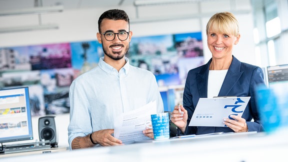 Eine Frau und ein Mann stehen in einem Großraumbüro und lächeln.