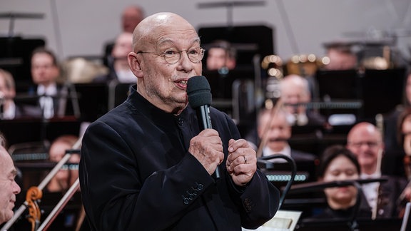 Dennis Russell Davies mit Mikrofon im Interview auf der Bühne des Leipziger Gewandhauses beim Festkonzert zum 100. Geburtstag der MDR-Ensembles