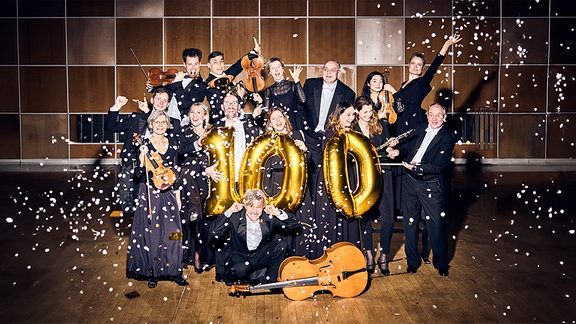 Mitglieder von MDR-Sinfonieorchester und MDR-Rundfunkchor in Konzertkleidung mit Instrumenten, Konfetti und einer goldenen 100 aus Folienballons.