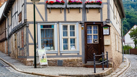 Ein zweigeschossiges Fachwerkhaus an einer Straßenecke, mit einer Straßenlaterne und Blumen vor den Fenstern. 