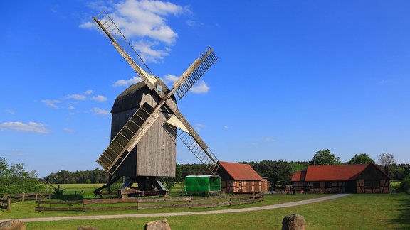 m Zentrum des Bildes ist eine gut erhaltene Bockwindmühle aus Holz zu sehen, sie steht vor einem hellgrünen Feld, dahinter ist Wald, rechts neben der Mühle befinden sich zwei Fachwerkgebäude mit roten Dächern.