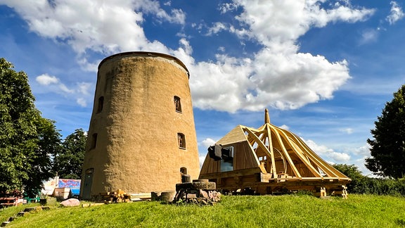 Das Bild zeigt den dicken, massiven, runden Steinturm der Düppler Mühle auf einer kleinen grünen Anhöhe, daneben liegt der hölzerne Dachstuhl für die neue Mühlenhaube.