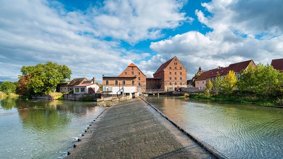 Im Vordergrund ein flaches Wehr im Fluss Unstrut, dahinter die historische Wassermühle mit den Giebelansichten der beiden drei- bzw. viergeschossigen zentralen Gebäude mit hohem Spitzdach und aus rotem Backstein sowie mehreren kleinen Nebengebäuden und Bäumen