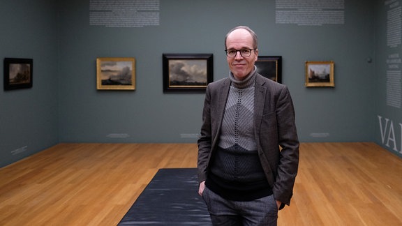 Zu sehen ist Stefan Weppelmann, Museumsleiter im Museum der bildenden Künste in Leipzig. Er steht in einem Raum, in dem mehrere Gemälde hängen und trägt eine schwarze Anzugjacke und eine Brille. Die Hände hat er in den Hosentaschen und er lächelt in die Kamera.