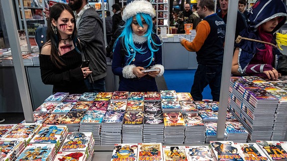 Auf einem Tisch liegen Naruto Manga Hefte. Dahinter stehen zwei interessierte Cosplayer.