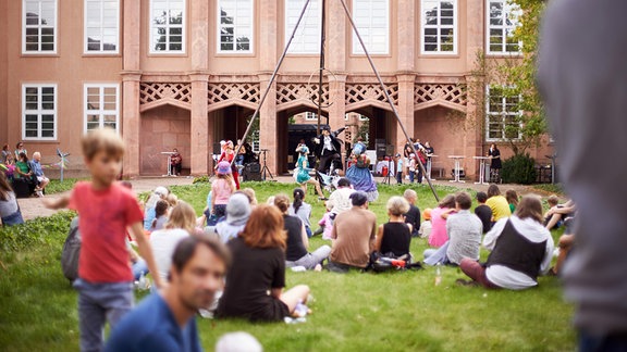 Personen sitzen auf der Wiese des Grassimuseums, eine Gruppe verkleideter Menschen zeigt eine Performance.