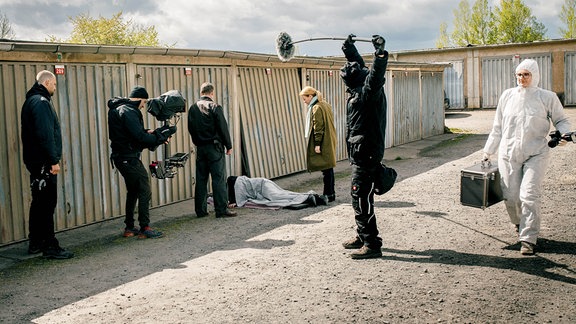 Sieben Personen sind auf einem Filmset an einer Reihe von Garagen zu sehen: Schauspielerinnen und Schauspieler sowie Mitglieder der Filmcrew mit Kamera und Tonangel.