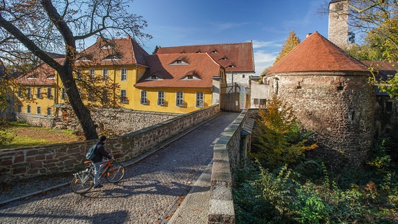 Blick über eine steinerne Brücke auf eine Burganlage mit mehreren Gebäuden, teils leuchtend gelb angestrichen, darüber blauer Himmel