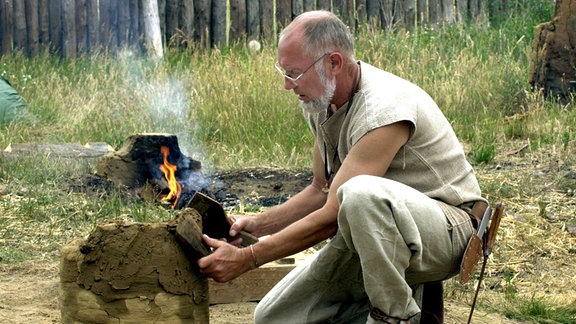 Ein Mann mit Brille und Backenbart kniet im Freien am Boden vor einem Feuer und hantiert mit Spatel und Lehm, um einen zylindrischen Ofen zu bauen.