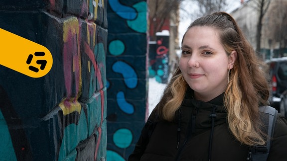 Eine junge Frau mit mittellangem, blondem Haar blickt seitlich in die Kamera und lächelt. Im Hintergrund eine Wand mit Graffiti.