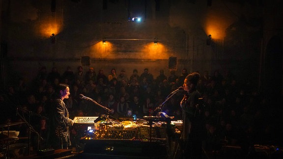 Die beiden Musikerinnen des Duos Olicia stehen sich bei einem Konzert auf der Bühne gegenüber. Im Hintergrund im schummrigen Licht sitzt das Publikum. 