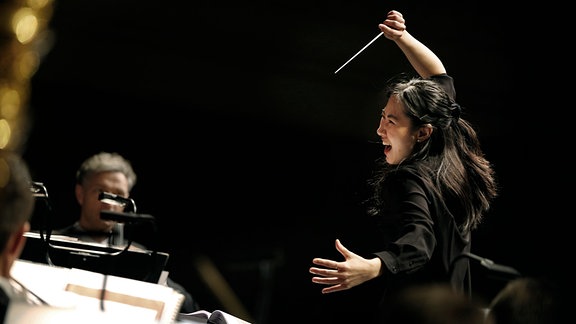 Eine Frau steht vor einem Orchester und schwingt wild den Taktstock über dem Kopf.