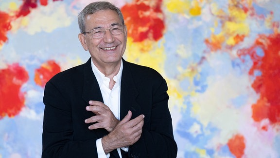 Orhan Pamuk, ein Mann mit grauen Haaren und einer Brille steht in einem Anzug vor einem farbigen Hintergrund. 