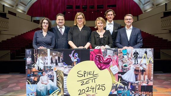 Sechs Personen stehen an einem Pult mit einer Bildcollage. Im Hintergrund ist der Publikumssaal eines Theaters zu erkennen.