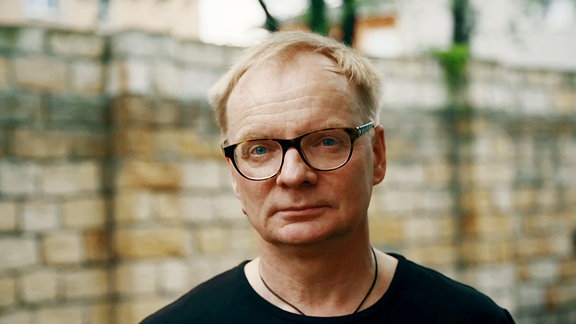 Uwe Steimle, ein Mann mit bloden Haaren, er trägt eine Brille mit schwarzem Rahmen und einen dunklen Pullover. 