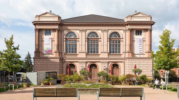 Landestheater Eisenach: Fasse eines großes hisorischen Gebäude aus Sandstein mit drei großen oben gebogenen Fenstern, davor ein Platz mit Pflanzen und zwei Bänken