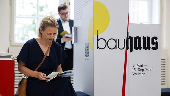 Eine Frau in dunkelblauem Kleid Sitz neben einem Aufsteller mit der Aufschrift "Bauhaus".