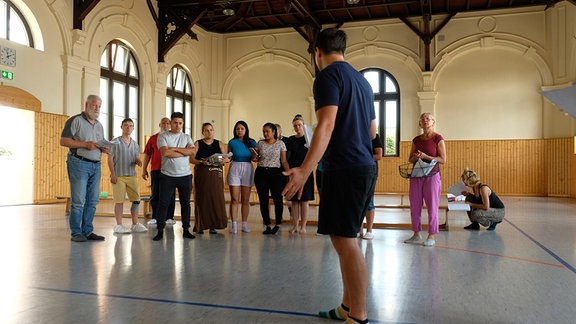 In einer Turnhalle steht ein Mann vor einer Gruppe weitere Menschen, eine Konfrontation spielend.