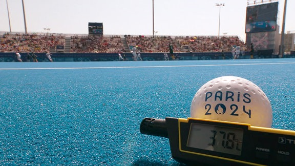 Ein Hockeyball liegt auf einem Hockeyfeld zusammen mit einem Thermometer, das 37,8 Grad Celsius anzeigt.