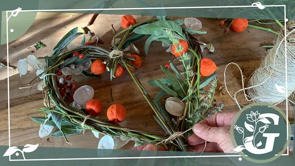 Ein Herz wurde aus Holztrieben gewunden. Zwei Hände befestigen Blätter und Früchte am Herz.