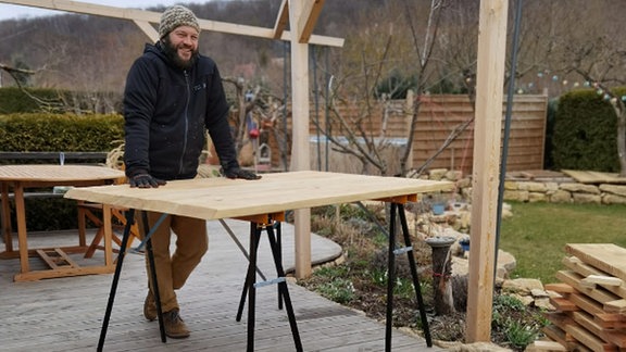 Auf ener Terrasse liegt viel Holz. Frank Krause steht an einem Arbeitstisch.