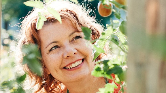 Eine Frau mit grüner Schürze steht in einem Garten vor einem Pfirsichbaum mit Früchten.  