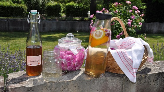 Auf einer Mauer stehen eine Flasche mit Sirup, ein Henkelglas mit Flüssigkeit, ein Glasgefäß mit Deckel und Rosen, eine Karaffe mit Früchten und Flüssigkeit sowie einem Körbchen.